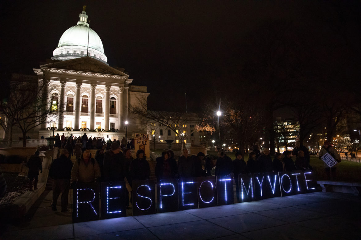 People gather at night outside a lit domed building with illuminated letters spelling “RESPECT MY VOTE” next to a sidewalk.