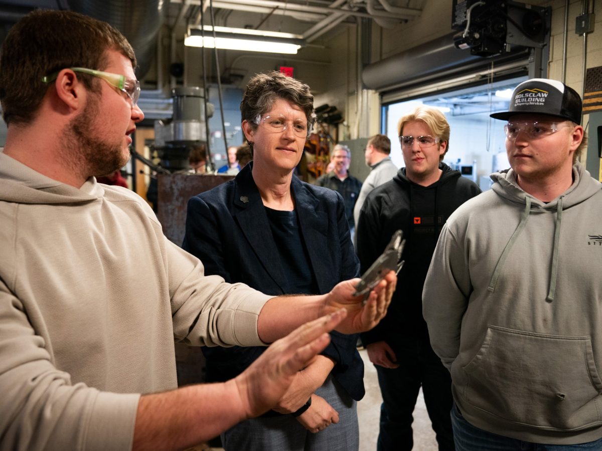 People wearing safety glasses stand in a workshop while one person holds a metal object and gestures.