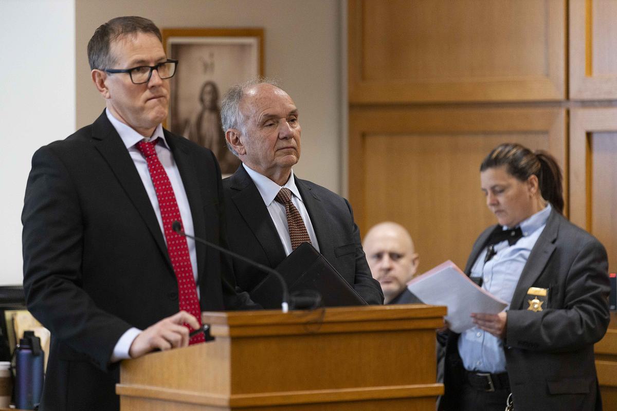 Two people in suits stand at a podium in a wood-paneled room, with another person nearby holding papers and wearing a badge on a jacket.