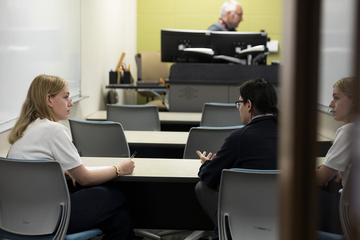 A woman sitting on the left side of a two-person desk takes notes while turning to a person sitting at the righthand side of the desk. An instructor sits at a desk at the front of the room.