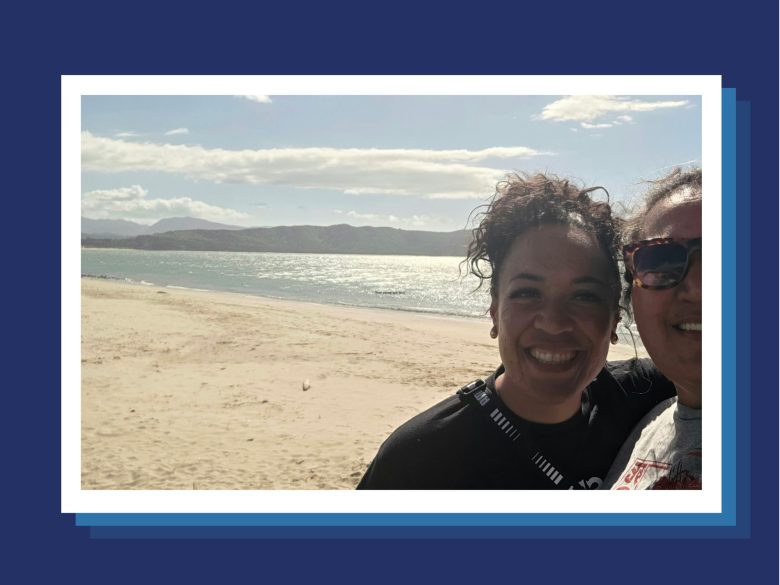 Two people smile for a selfie on a sandy beach with water, hills and clouds visible in the background.
