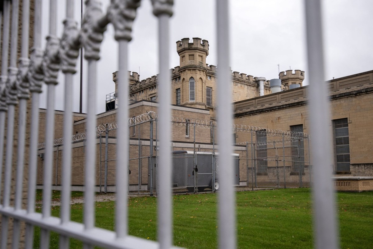 Metal fence in foreground with view of a tan brick building topped with guard towers, barred windows and fencing lined with razor wire under an overcast sky